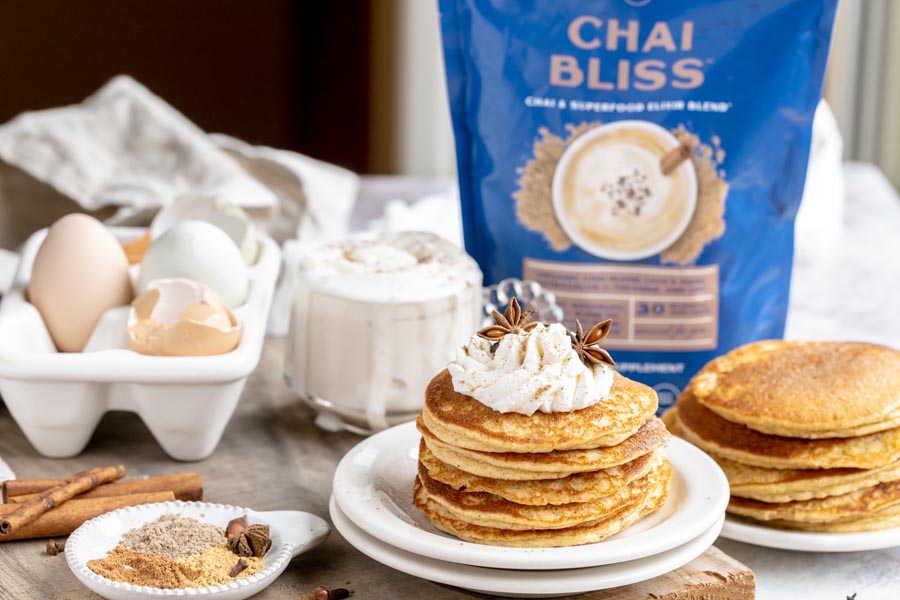 a bag of chai bliss sits behind two stacks of pancakes; spices, a latte and eggs sit nearby