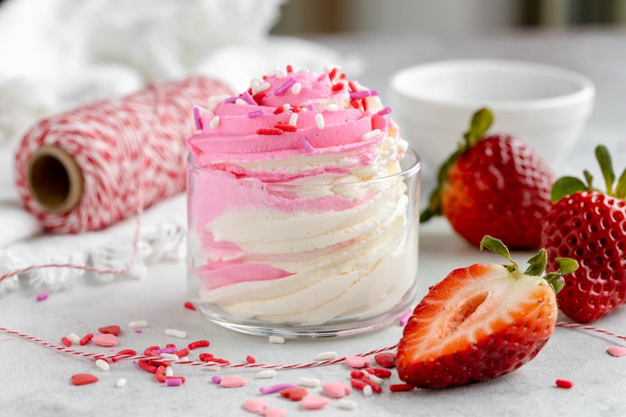 creamy cheesecake fluff in a clear bowl white and pink colored topped with sprinkles and strawberries lie nearby