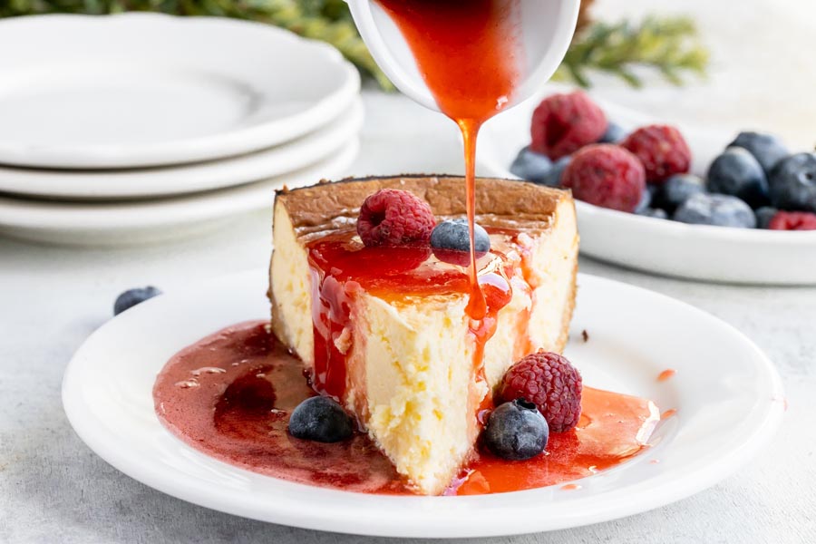pouring red strawberry sauce over a slice of cheesecake