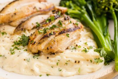 Grilled chicken on top of creamy fettucine alfredo next to broccoli.