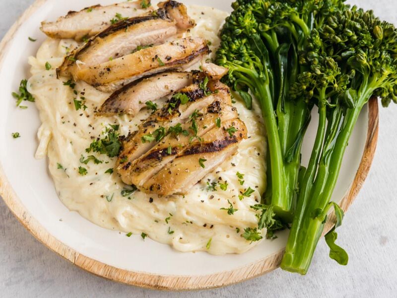A plate of chicken alfredo next to cooked broccolini.