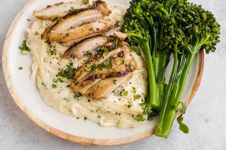 A plate of chicken alfredo next to cooked broccolini.
