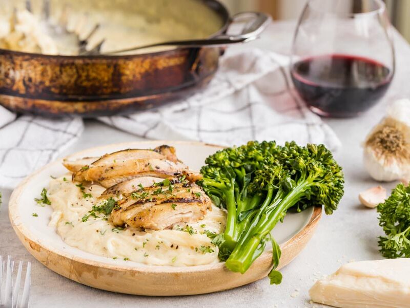 A dinner plate filled with creamy white chicken alfredo and broccoli. With a glass of wine in the background.