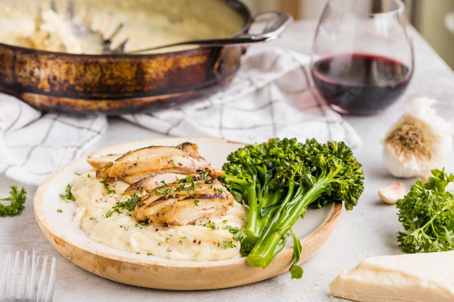 A dinner plate filled with creamy white chicken alfredo and broccoli. With a glass of wine in the background.