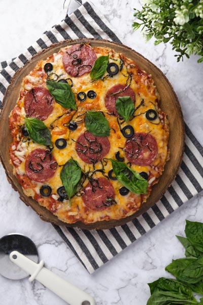 Unsliced whole pizza on a pizza stone over a towel. Pizza is topped with basil leaves, sliced olive and pepperoni.
