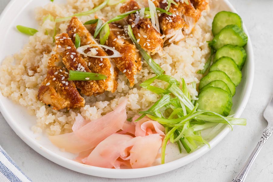 Close up of sesame and panko crusted chicken over cauliflower rice and topped with sliced onion.