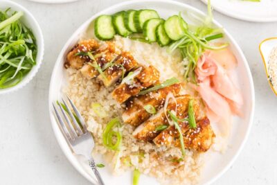 A plate with katsu chicken over cauliflower rice next to sliced cucumber and pickled ginger.