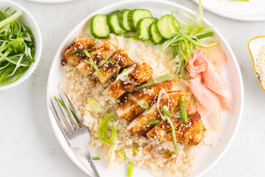 A plate with katsu chicken over cauliflower rice next to sliced cucumber and pickled ginger.