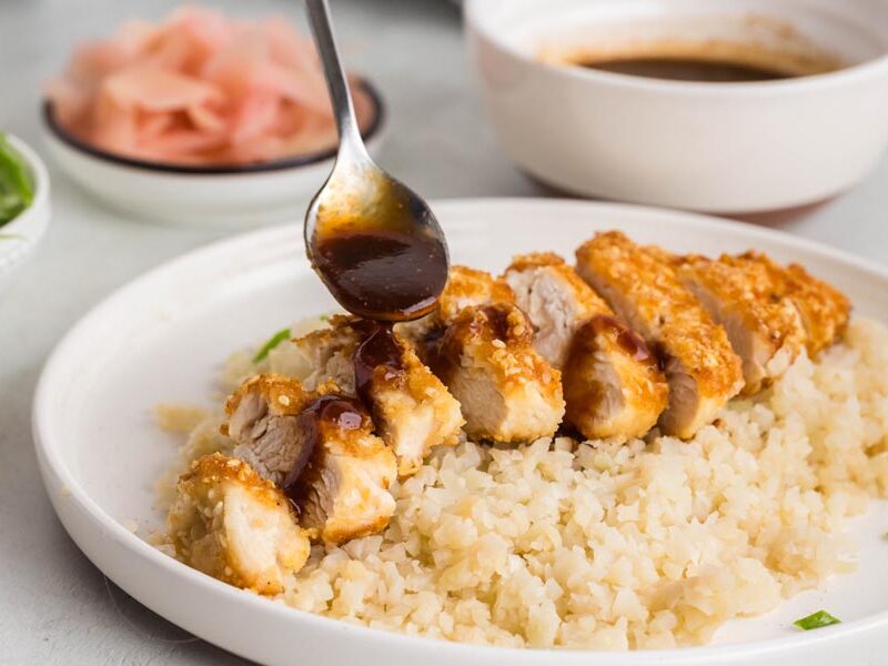 A spoon pouring tonkatsu sauce over breaded chicken sitting on rice.