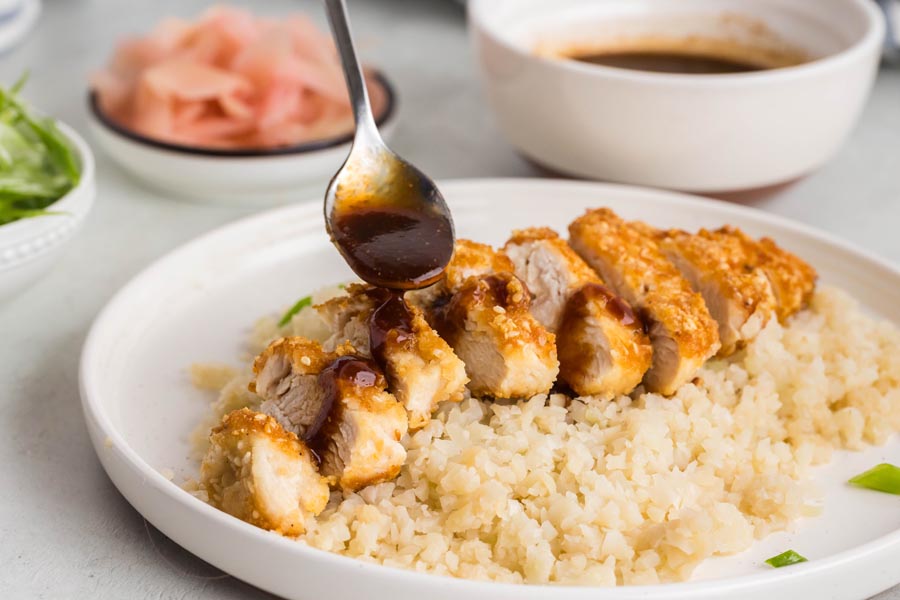A spoon pouring tonkatsu sauce over breaded chicken sitting on rice.