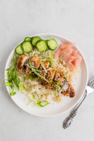 A plate with chicken katsu topped with sauce, sesame seeds and sliced green onions next to sliced cucumbers and ginger.