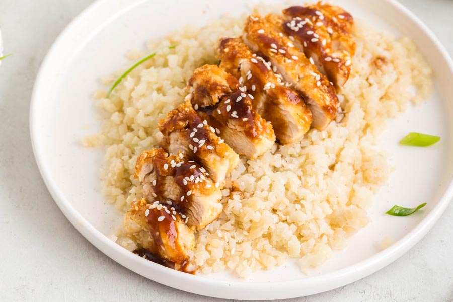 Sliced keto katsu chicken over a bead of cauliflower rice on a white plate and topped with sauce and sesame seeds.