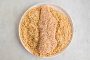 A chicken breast coated with pork panko and sesame seeds on a plate with more breading.