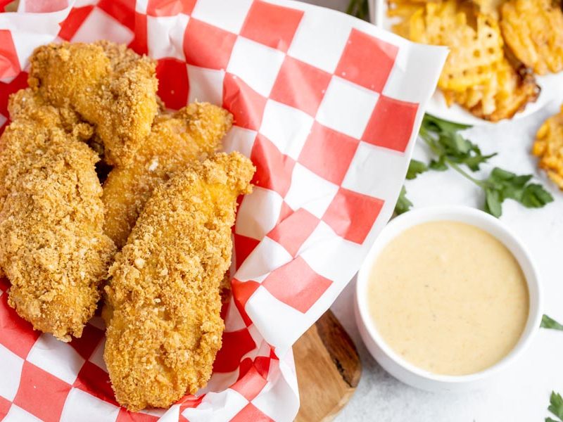 Breaded chicken tenders in a checkered basket with dipping sauce and waffle fries.