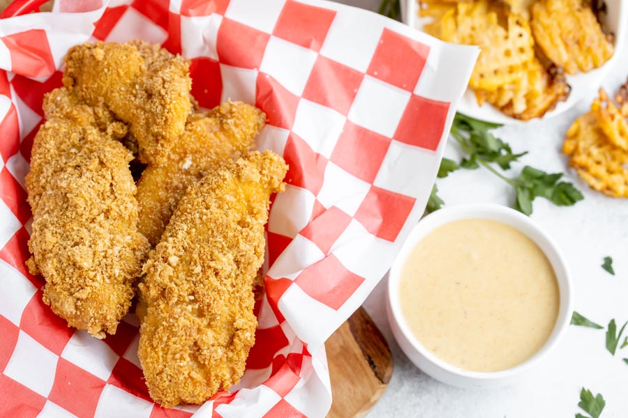 a basket of chicken strips in a red and white checkers parchment paper basket next to dipping sauce and crispy waffle fries