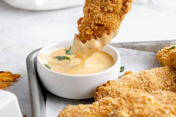 dipping a crunch chicken strip in a white bowl filled with honey mustard sauce