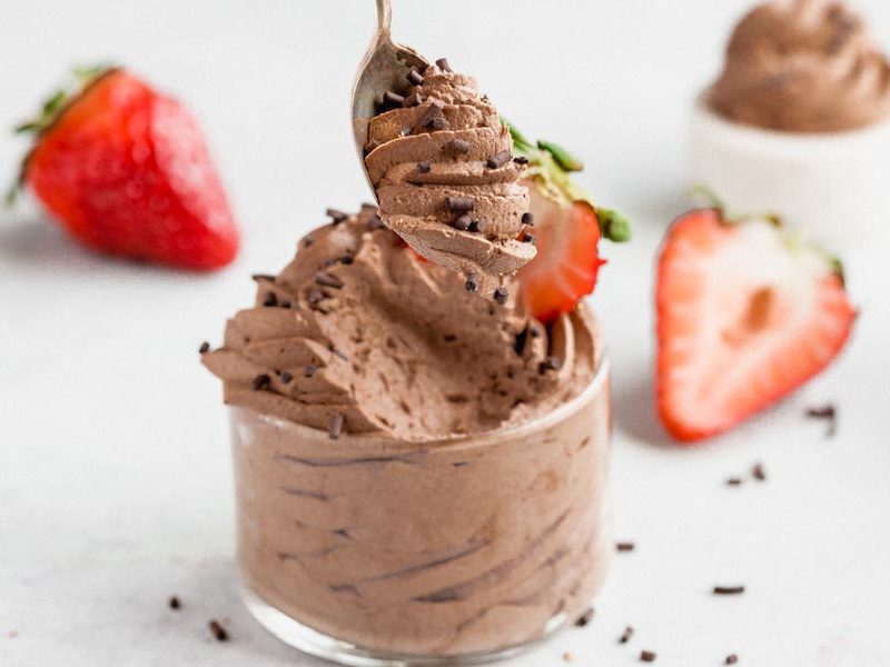 Spoonful of keto chocolate mousse above a glass dish with strawberries and chocolate sprinkles.