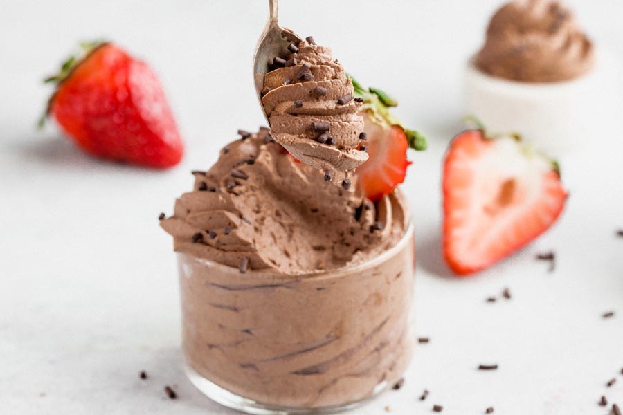a spoonful of creamy chocolate mousse in front of a dish of it while chocolate sprinkles are tossed nearby and slice strawberries lay in the background