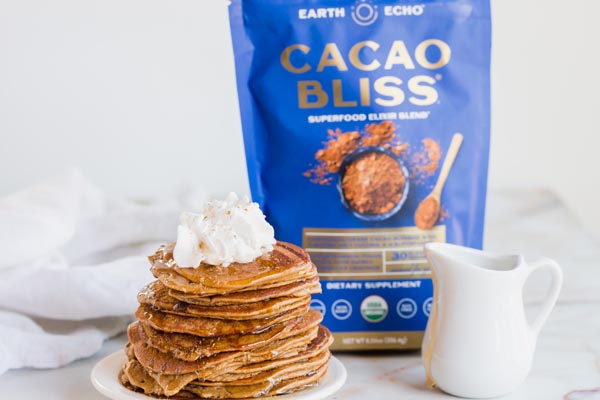 a stack of chocolate pancakes in front of a blue bag of cacao bliss and syrup