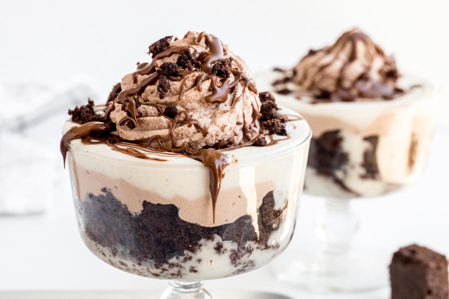 Melted chocolate dripping down the side of a trifle dish containing a chocolate cake pudding trifle topped with chocolate whipped cream, crumbled cake and melted chocolate.