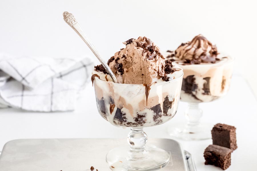 A spoon sitting in a small trifle bowl with layers of chocolate cake and cream. Chunks of chocolate cake lay beside.