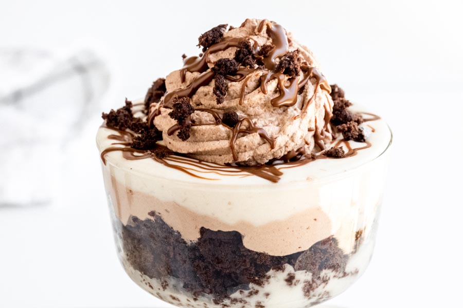 A small dish containing layers of white pudding, chocolate cream and chocolate cake. A swirl of chocolate cream is on top and is topped with crumbled cake and melted chocolate.