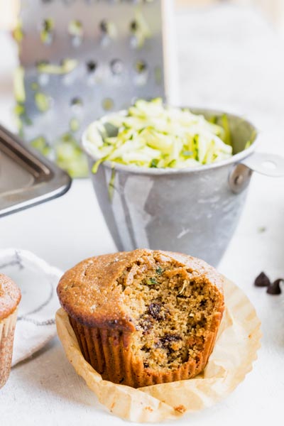 a muffin with a big bite taken out of it and a measuring cup filled with zucchini behind it