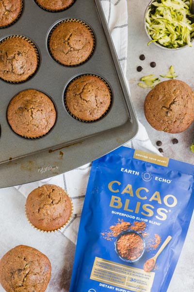 bakery style muffins in a muffin tin with shredded zucchini near and a bag of cacao bliss