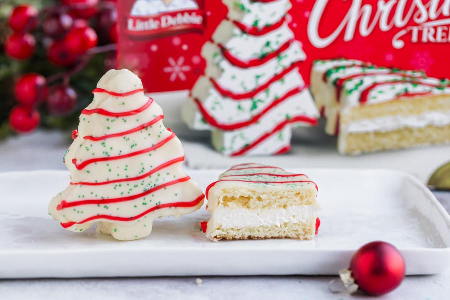 A white cake in the shape of a Christmas tree stands on it's end next to a half a cake cut in half to show the creamy center in front of a box of Little Debbie's Christmas Tree Cakes.