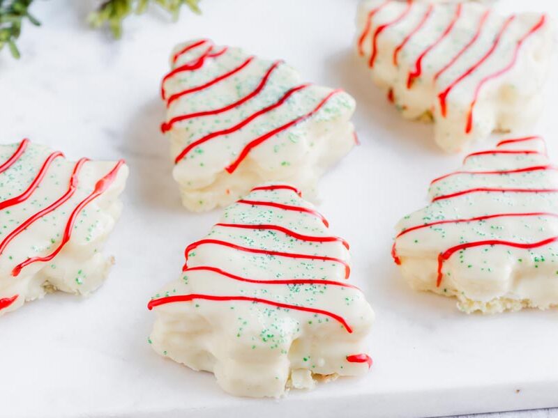 Five snack cakes on a marble slab in the shape of christmas trees and covered with white chocolate, green sprinkles and red stripes.