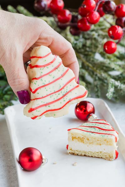 A hand holding onto the a small snack cake shaped like a Christmas tree over a plate with another cake that is cut in half to show the inside layers of white cake and creamy middle filling.