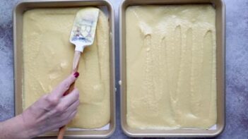 A hand spreading cake batter onto two quarter sheet baking pans with a spatula.