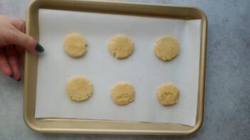 A hand holding a baking sheet with round unbaked cookies on it.