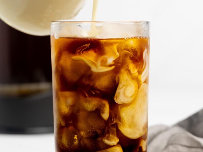 Homemade keto coffee creamer being poured into a glass of iced coffee over ice cubes.