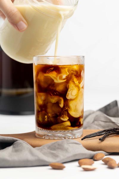 Keto coffee creamer being poured into iced coffee with vanilla beans and almonds arranged nearby.