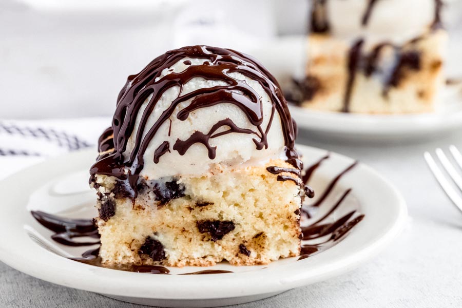 A cookie bar on a white plate topped with a scoop of ice cream and chocolate sauce.