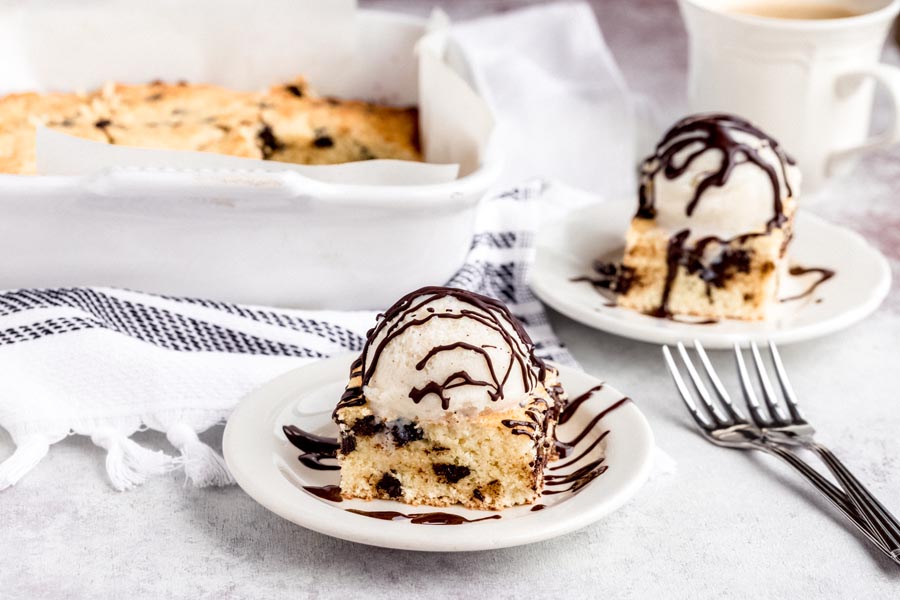 Two plates of cookie bars a la mode and covered in chocolate with a pan of cookie bars in the background and two forks.