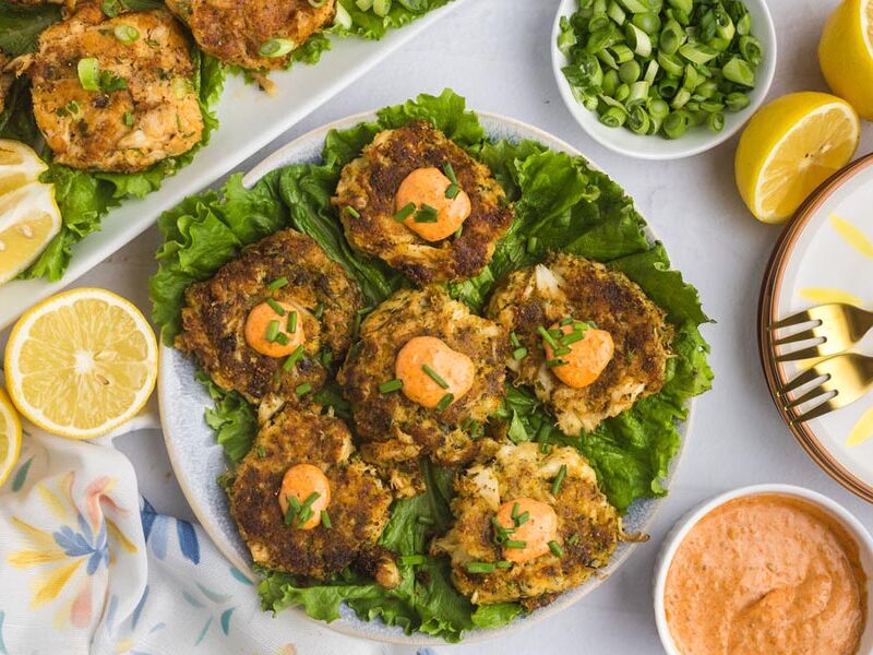A platter of crab cakes topped with sauce next to fresh lemon and green onions.