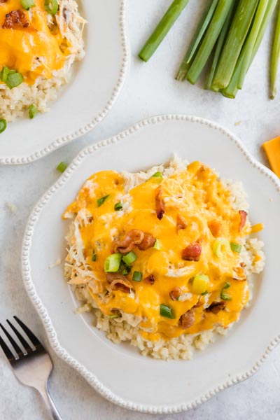 a plate with crack chicken on riced cauliflower and topped with melted cheese, bacon and green onion. Next to a bundle of fresh green onions.