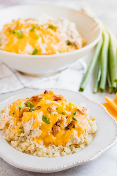 A plate with creamy crack chicken on top of rice with a bowl of more cheesy chicken before and a bundle of green onions.
