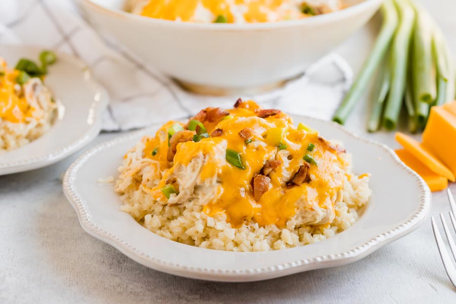 A plate of cheesy crack chicken and bacon over cauli rice next to sliced cheddar and a bundle of green onions.