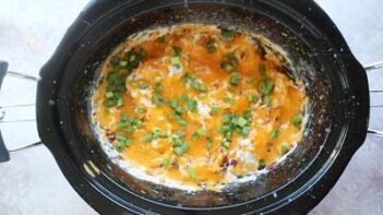 A crock pot with finished crack chicken inside Melted cheddar cheese is one top with sliced green onions sprinkled throughout.