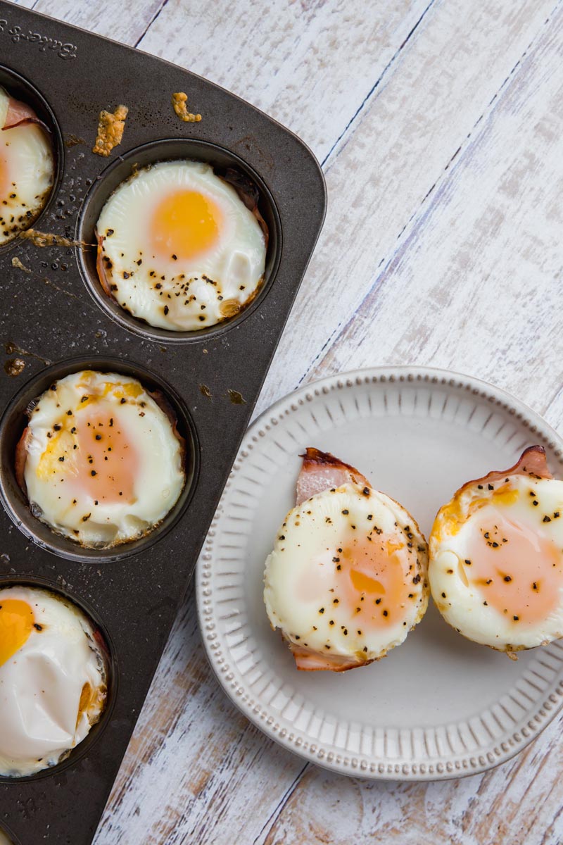 two egg cups on a plate with a full row of baked egg cups in a tray next to it