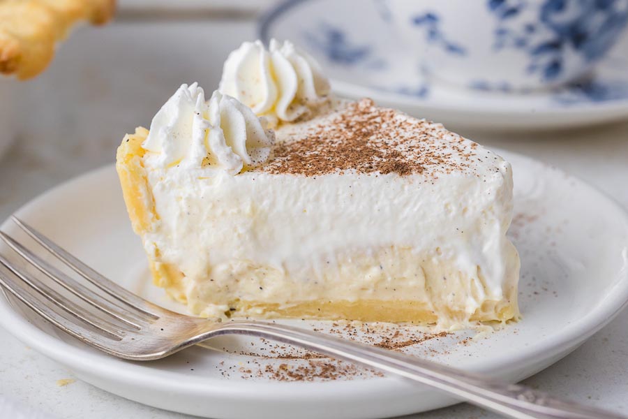 A slice of creamy keto eggnog pie topped with whipped cream and a dusting of nutmeg sits on a white plate with a fork.