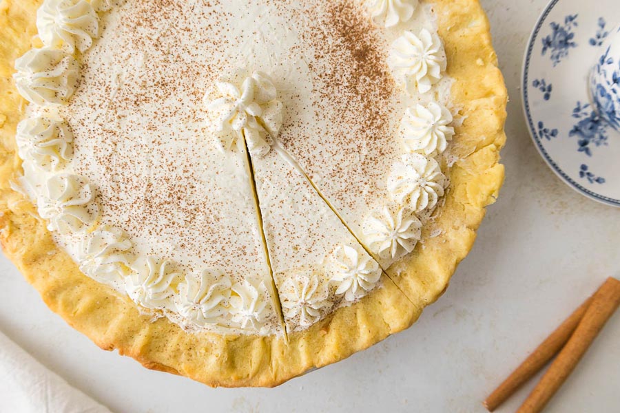 An overhead view of a full keto eggnog pie with a golden crust, piped whipped cream border, and a slice cut out.