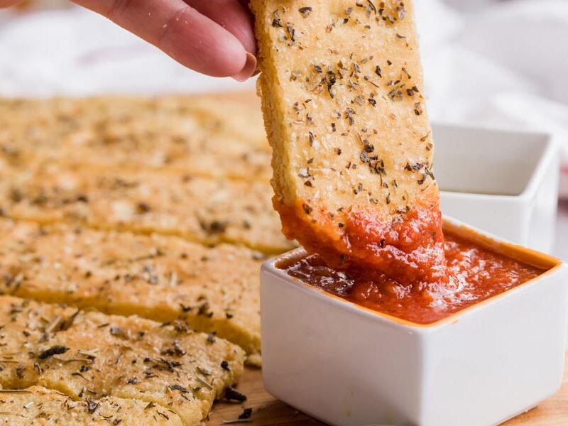 A hand dipping a piece of focaccia bread into a bowl of marinara sauce.