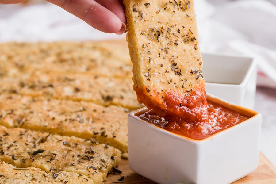 A hand dipping a piece of focaccia bread into a bowl of marinara sauce.