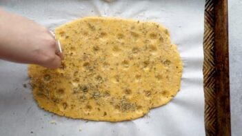 Unbaked focaccia with dimples and a hand sprinkles fresh herbs all over.