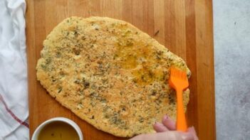 A hand brushing olive oil on focaccia bread with an orange silicone brush.