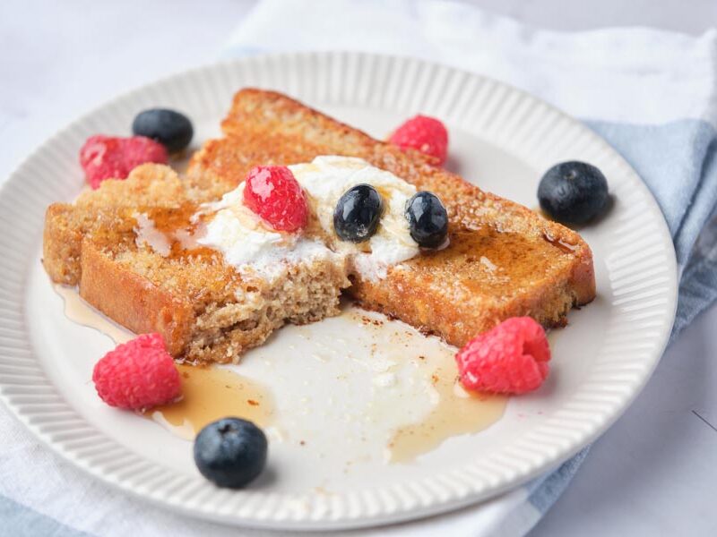 A bite taken out of a slice of keto french toast that is topped with whipped cream, berries and syrup.